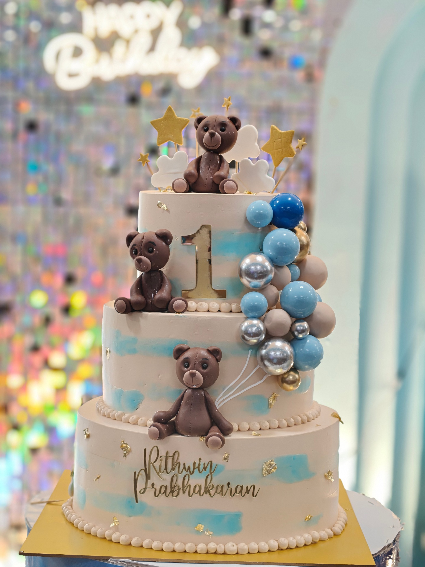 Three-tier teddy bear themed first birthday cake with blue balloons and gold accents